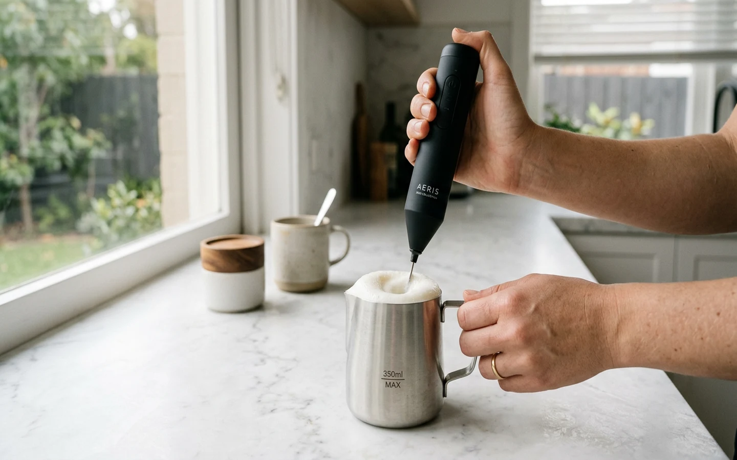 Electric milk frother creating frothy milk in a stainless steel jug on a kitchen bench